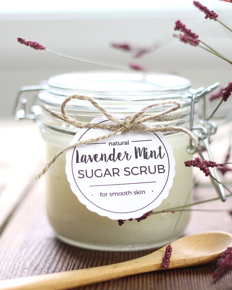 A clear jar filled with natural lavender mint sugar scrub is sealed with a metal clasp and decorative twine. The label reads "natural Lavender Mint SUGAR SCRUB for smooth skin." Dried lavender sprigs and a wooden spoon are placed artistically around the jar, making it one of the perfect essential oil gift ideas.