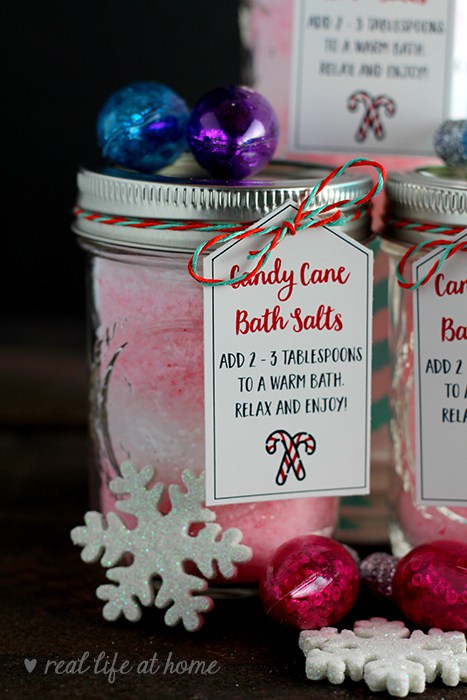 Glass jars filled with pink candy cane bath salts, topped with colorful ornaments and labeled with usage instructions, make charming essential oil gift ideas. Adorned with red and white twine, they are accompanied by decorative snowflakes and shiny baubles on a dark background.