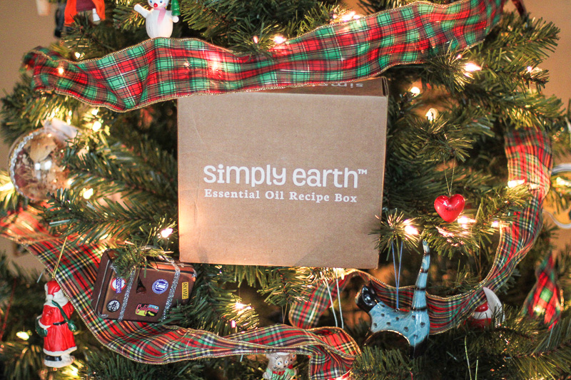 A Christmas tree adorned with colorful ornaments and a plaid ribbon features a prominently placed brown box with the text "simply earth™ Essential Oil Recipe Box," offering perfect essential oil gift ideas. The tree is lit with warm white lights and various festive decorations.