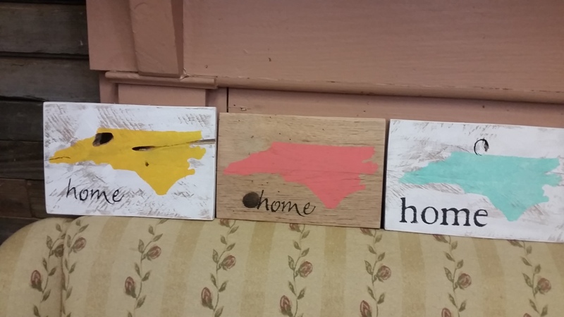 Displayed on a floral-patterned couch, these three wooden plaques featuring shapes resembling the state of North Carolina are part of the hottest crafts trend. Painted in yellow, coral, and teal from left to right, each plaque reads "home" at the bottom. Perfect for those who make and sell handmade goods!