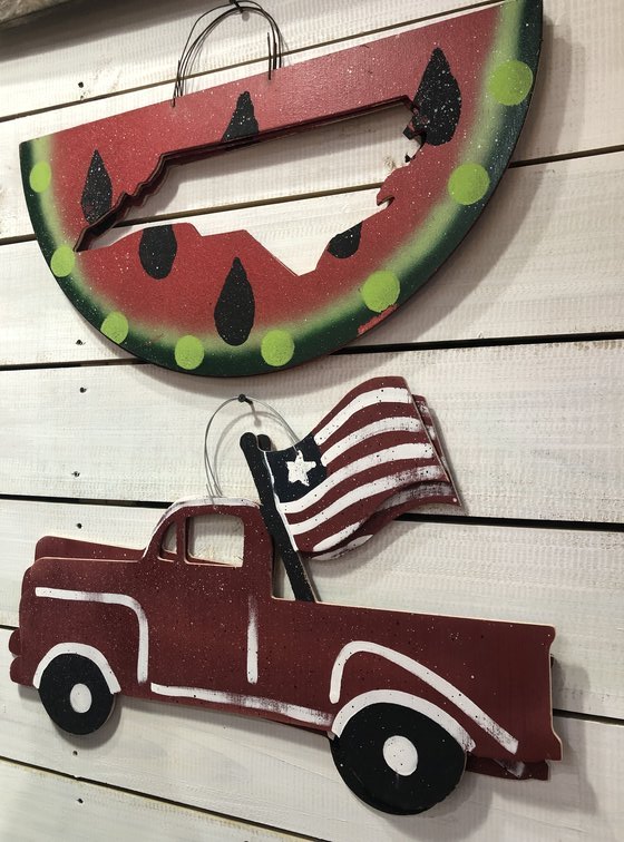 A decorative wall display from the hottest crafts features a rustic metal cut-out of a watermelon slice above a red vintage truck holding an American flag, both hanging on a light wooden background. Perfect for those looking to make and sell unique decor.