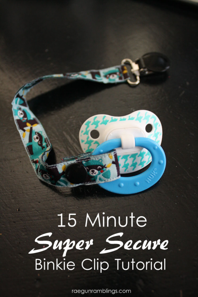 A pacifier with a blue ring is attached to a fabric strap featuring a penguin design. The strap has a black clip at the end. Text at the bottom reads: "15 Minute Super Secure Binkie Clip Tutorial - Baby Craft Ideas.
