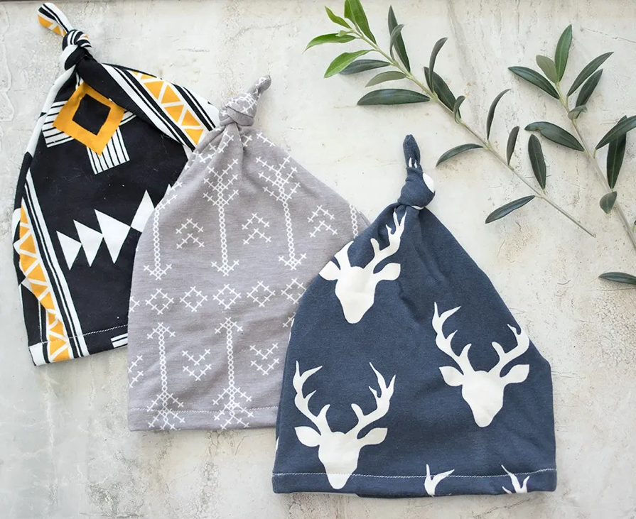 Three knotted baby hats arranged on a light surface next to some green leaves—perfect for 30+ baby craft ideas. The hats have different designs: one black with geometric shapes in white, yellow, and orange; one gray with white arrow patterns; and one blue with white deer head prints.