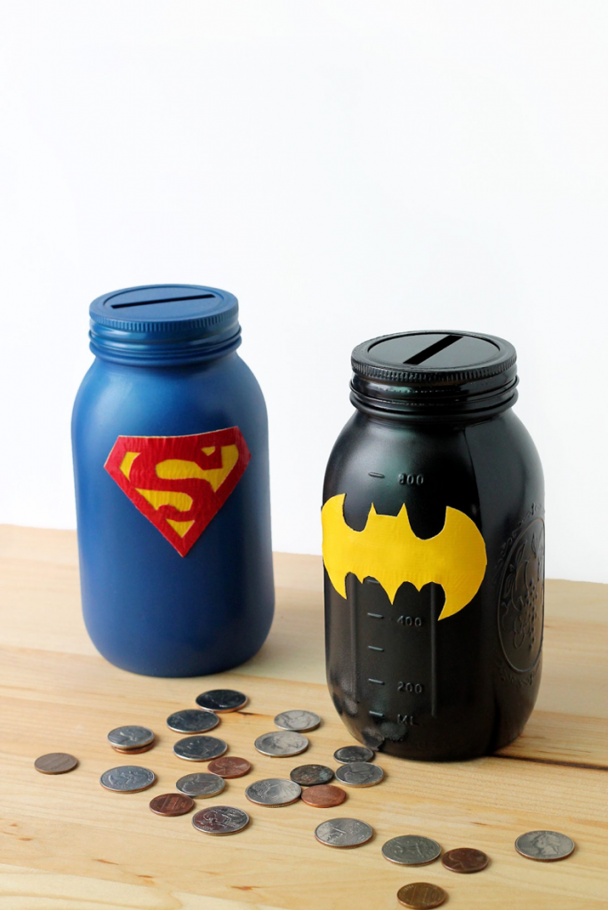 Two painted mason jars serve as coin banks, perfect for baby craft ideas. One jar is blue with a red and yellow Superman logo, while the other is black with a yellow Batman logo. Coins are scattered around the jars on a wooden surface, ready to inspire your next make and sell project. The background is plain white.