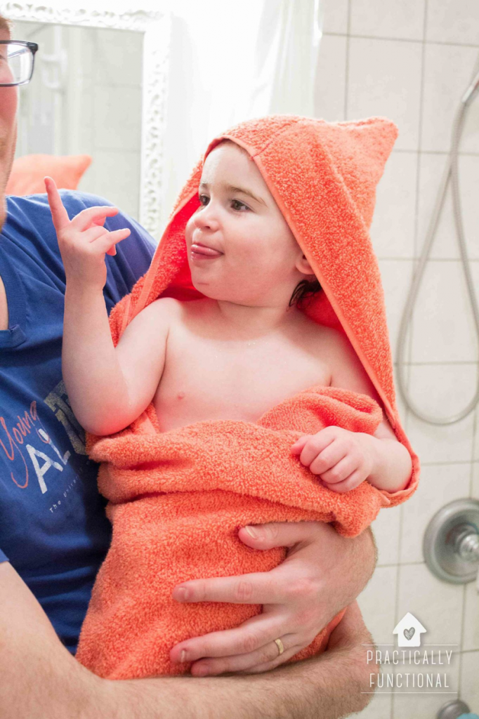 A small child with wet hair is wrapped in an orange towel with a hood, sticking out their tongue and making a playful hand gesture. An adult, partially visible on the left, is holding the child in a bathroom setting, showcasing one of those adorable baby craft ideas perfect for playful moments.