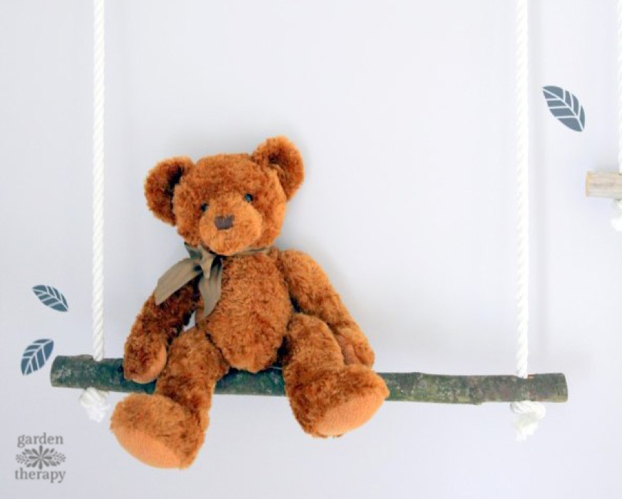 A brown teddy bear with a ribbon around its neck sits on a wooden swing made from a tree branch, hung by white ropes. The background is light-colored with a few blue leaf decals. Perfect for baby craft ideas or even to make and sell, the "Garden Therapy" logo is visible in the bottom left corner.