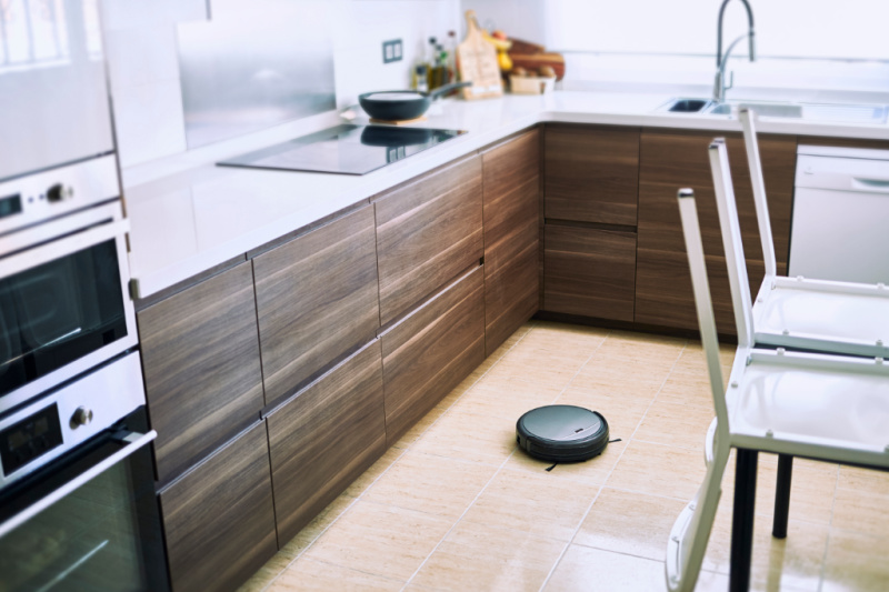 A modern kitchen with brown wooden cabinets, a built-in oven and microwave, an induction cooktop, and a countertop with utensils and bottles. In the center of the tiled floor, a robotic vacuum cleaner operates near a chair, making for an easy-to-stick-to cleaning routine.