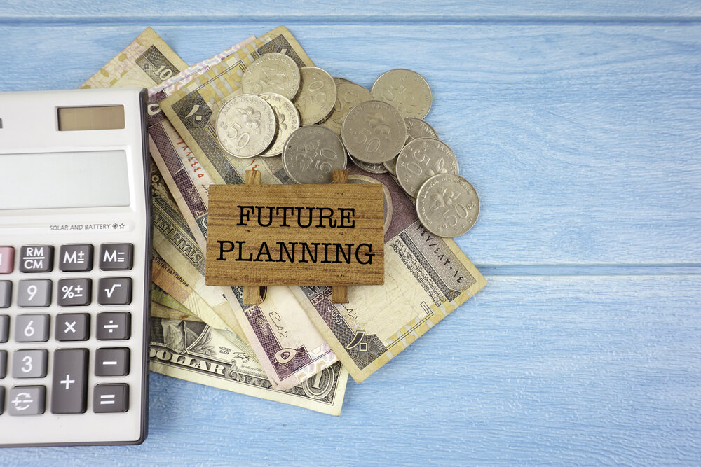 A calculator sits next to piles of various currencies, including coins and paper bills. Among the money, a small wooden sign reads "FUTURE PLANNING," emphasizing the importance of managing finances. The background is a light blue wooden surface.