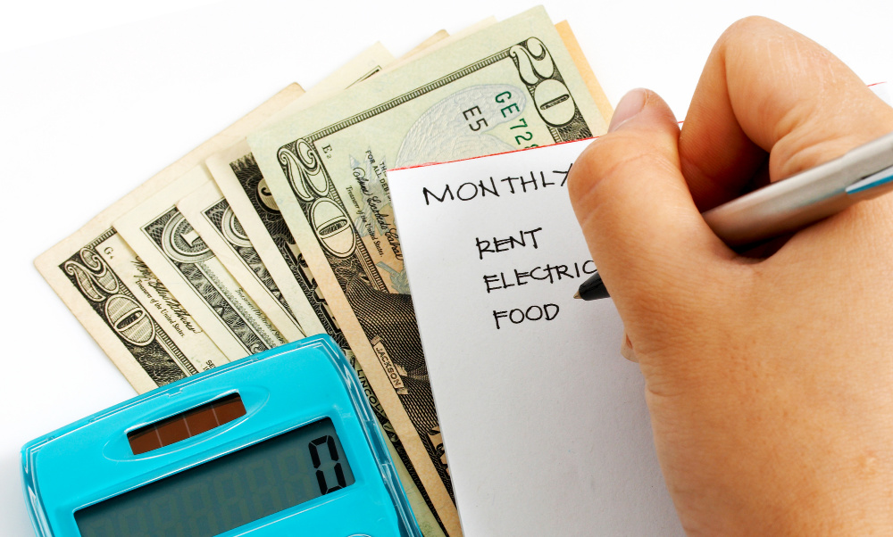 A hand writes a monthly budget list with categories for rent, electric, and food on a notepad. Several U.S. dollar bills and a blue calculator, showing zero, lie next to the notepad on a white surface—an essential exercise in personal finance during uncertain times.