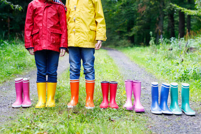 7 pairs of rainboots in the colors of the rainbow sitting on a gravel road. Two little girls are wearing 2 of the pairs. 