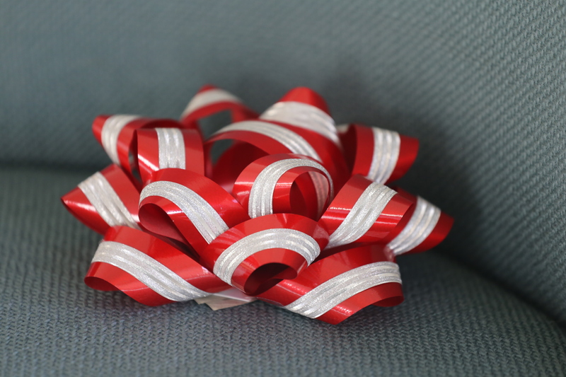 A brilliant decorative red ribbon bow with white stripes rests on a gray textured fabric surface. The bow, perfect for Dollar Tree money gifts, has multiple loops creating a festive and elegant appearance.