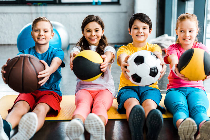 4 little kids holding different sports balls in front of them. 
