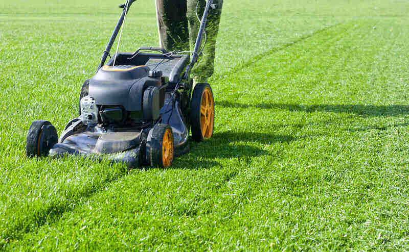 A person is pushing a lawnmower with orange wheels across a well-manicured, green lawn. The mower is in motion, creating clear, parallel lines of cut grass. The scene is sunlit, showcasing the vibrant color of the grass—an idyllic way to make $500 extra per month.