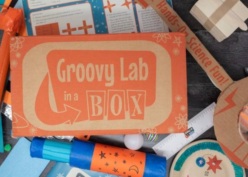 A brown cardboard box that has the words Groovy Lab in a BOX on it. It is sitting on top of science type decorations.