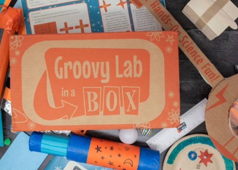 A brown cardboard box that has the words Groovy Lab in a BOX on it. It is sitting on top of science type decorations.