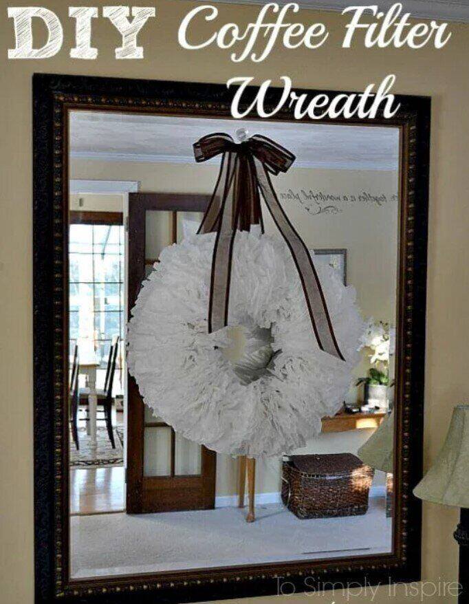 A large DIY wreath made from white coffee filters is hanging on a mirror, showcasing farmhouse charm. Topped with a brown ribbon bow, its reflection reveals a cozy, sunlit room with a wicker basket and lamp. This charming piece could easily grace flea markets or inspire your next style ventures.