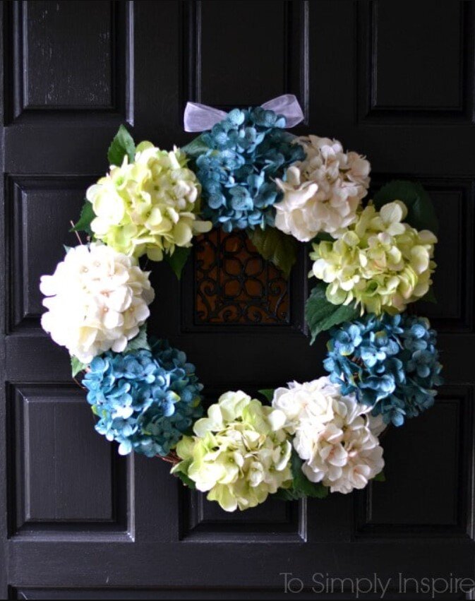A charming wreath made of green, blue, and white hydrangeas hangs on a dark wooden door, reminiscent of the delightful finds at farmhouse crafts fairs and flea markets.