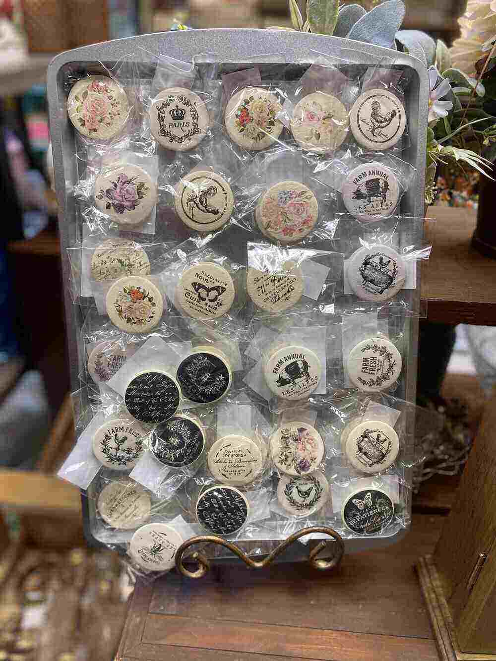 A display tray features various round buttons and magnets, each wrapped in clear packaging and perfect for crafts enthusiasts. The soaps are decorated with floral patterns, animal designs, and typography. Some depict vintage-style graphics, while others have rustic farm themes, making them the hottest items to make and sell.