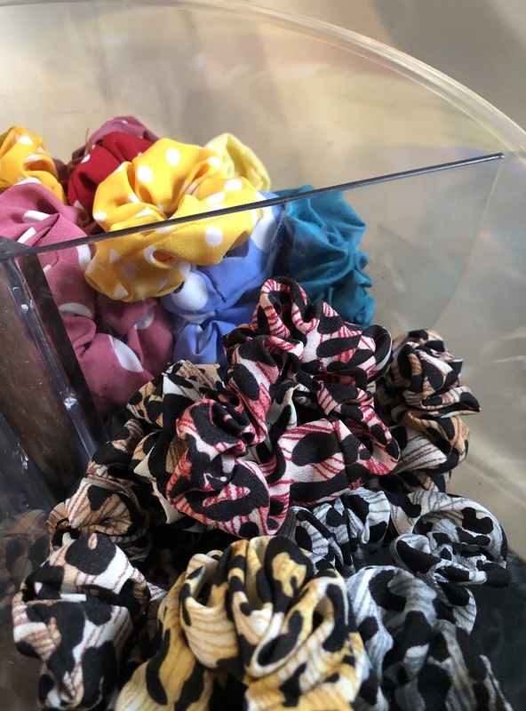 A clear container holds colorful fabric scrunchies, one of the hottest crafts to make and sell. Visible scrunchies include a yellow one with white polka dots, a pink one, a blue one, and several with intricate black, red, and beige patterns.