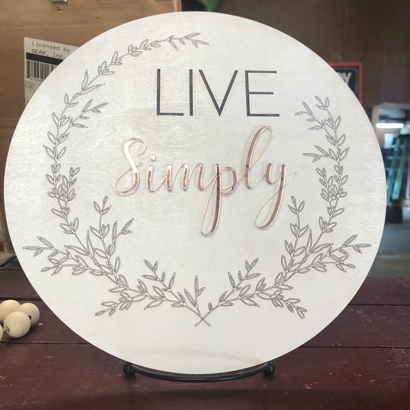A round white sign with the words "LIVE Simply" in the center. "LIVE" is written in black uppercase letters, and "Simply" is in a cursive font with a gold tone. The sign, one of the hottest crafts to make and sell, is decorated with a delicate wreath of leaves around the edges. It rests on a stand.