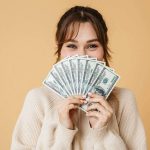 woman with brown hair holding money that is fanned out in front of her face.