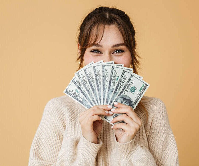 woman with brown hair holding money that is fanned out in front of her face.