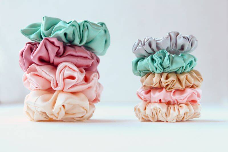 A close-up image of two stacks of colorful scrunchies, perfect as Easy Market Day Projects or Make and Sell items, on a white background. The left stack has five scrunchies in shades of green, pink, and beige. The right stack has four scrunchies in shades of gray, green, beige, and pink.
