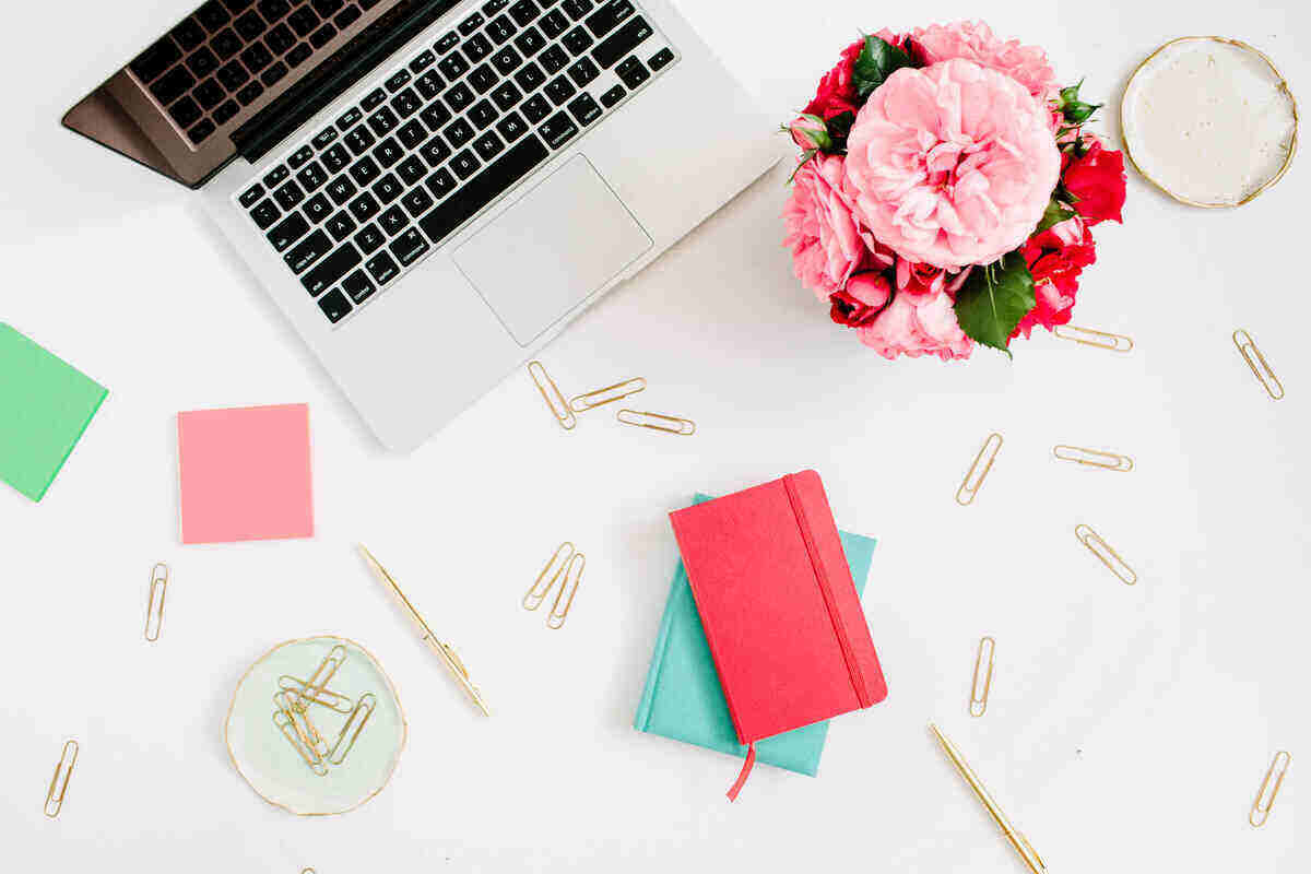 A tidy workspace features a laptop, a red notebook on top of a green notebook, scattered gold paperclips, a pink pen, pink and green sticky notes, a plate of paperclips, and a vase of pink flowers—perfect for brainstorming legit ways to make money online—all arranged on a white surface.