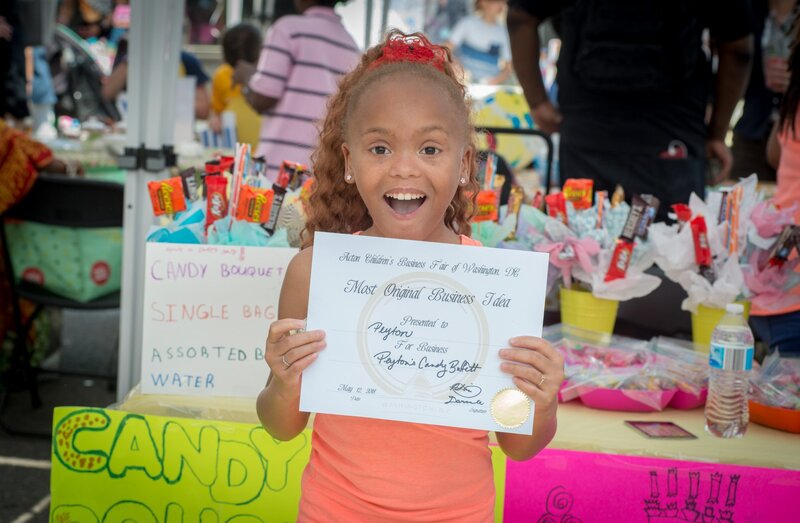 A smiling young girl with curly hair and a red bow holds up a certificate in front of her. The certificate reads "Most Original Business Talent" and is related to candy. Behind her, at the Children's Business Fair, is a booth displaying colorful candy bouquets and assorted candy.