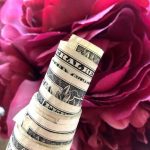 A close-up image of a rolled-up stack of U.S. dollar bills in front of vibrant pink flowers showcases one of the most unique ways to give money gifts. The bills are tightly wound, with the top bill showing part of "United States" and "America." The flowers are large, with delicate, layered petals.