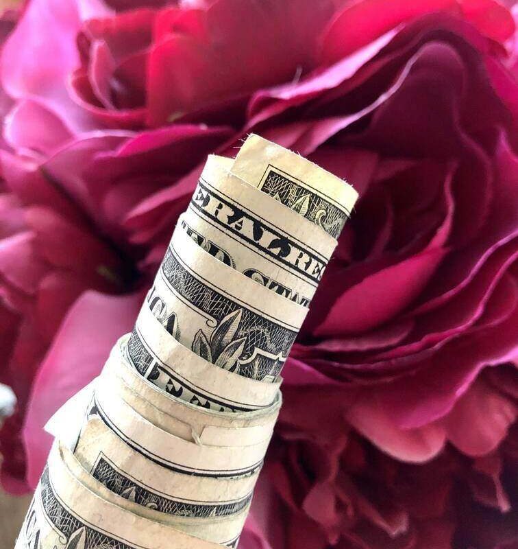 A close-up image of a rolled-up stack of U.S. dollar bills in front of vibrant pink flowers showcases one of the most unique ways to give money gifts. The bills are tightly wound, with the top bill showing part of "United States" and "America." The flowers are large, with delicate, layered petals.