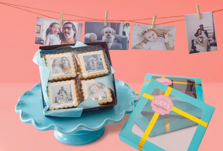 A blue dessert stand holds a box of cookies with printed photos on them. Behind it, a clothesline with family photos is clipped against a pink background. Another cookie box, open and tied with a yellow ribbon, sits next to the stand—perfect gifts for expectant grandparents.