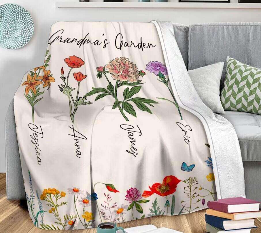 A white blanket with floral designs labelled "Grandma's Garden" lies on a couch. Each flower is associated with a name: Jessica, Anna, James, and Eric. Additional flowers and butterflies grace the bottom of the blanket. With cushions and a side table nearby, it makes perfect gifts for expectant grandparents.