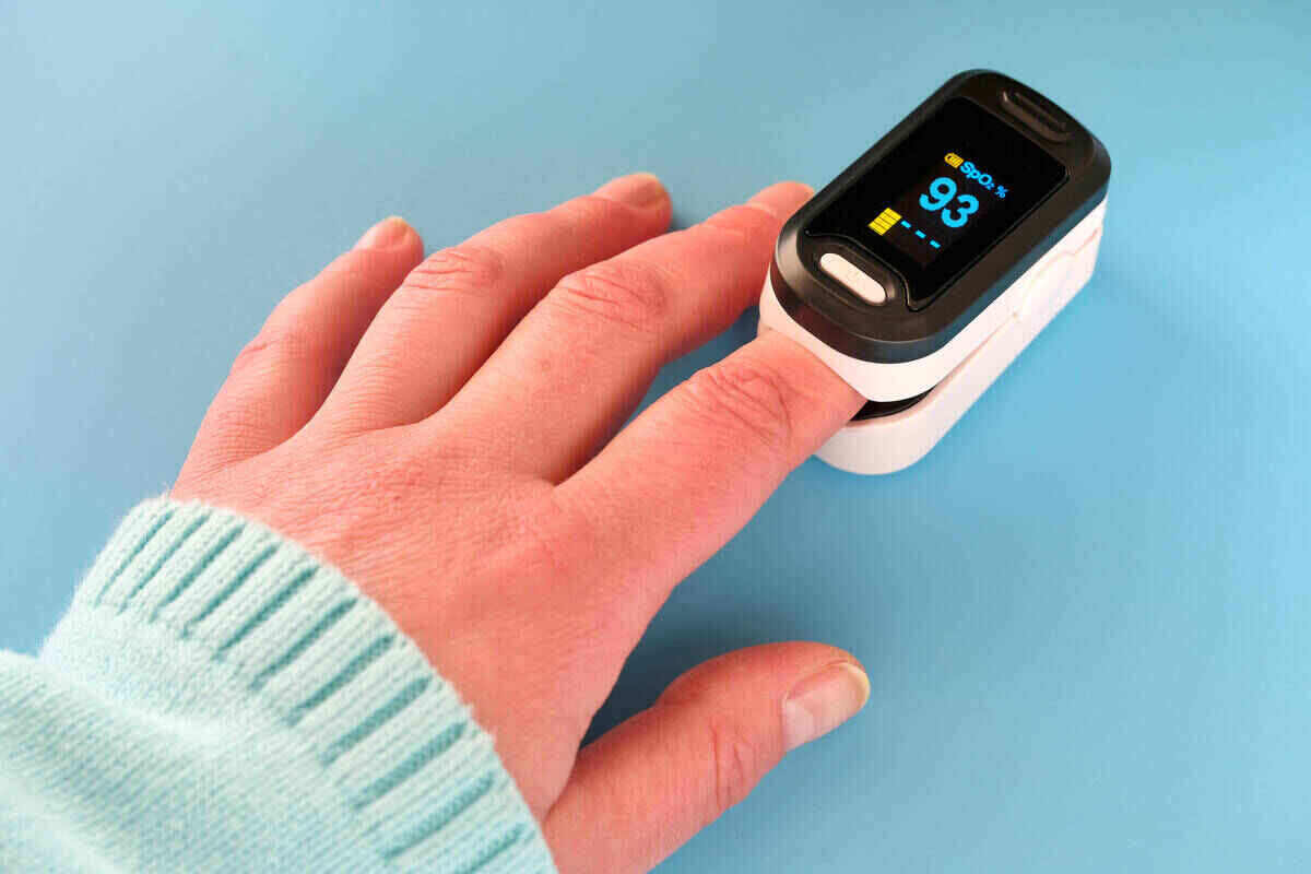 A hand with a light blue sleeve uses a pulse oximeter on the index finger. The item displays a reading of 93% oxygen saturation and a pulse rate of 74 beats per minute against a solid light blue background. This simple device has been critical in monitoring health, especially during Covid times.