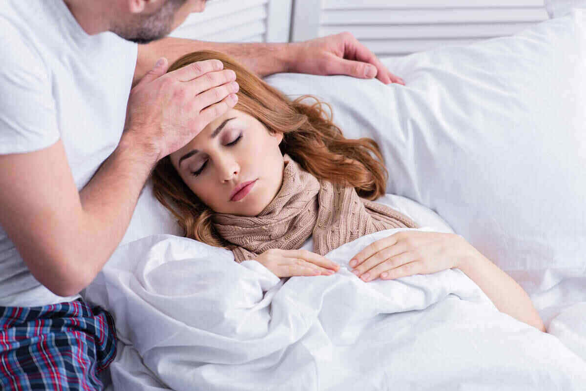 A woman lies in bed with a scarf around her neck, eyes closed, appearing unwell. A man sits beside her, gently touching her forehead as if checking for a fever. They are in a white, cozy bedroom setting with the woman covered in a blanket; it seems like they are battling Covid together.