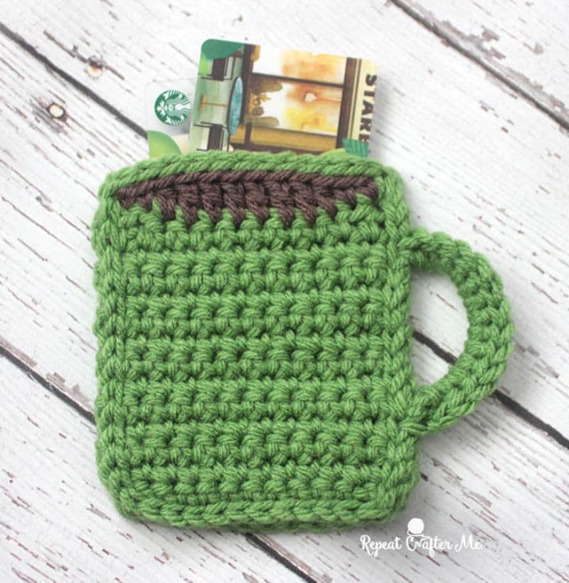 A small, green, hand-crocheted coffee mug with a handle is placed on a wooden surface. It serves as a cozy holder for a Starbucks gift card peeking out the top—a perfect choice for small gifts to friends. The mug features decorative brown stitching near the rim and bears the subtle watermark logo "Repeat Crafter Me.