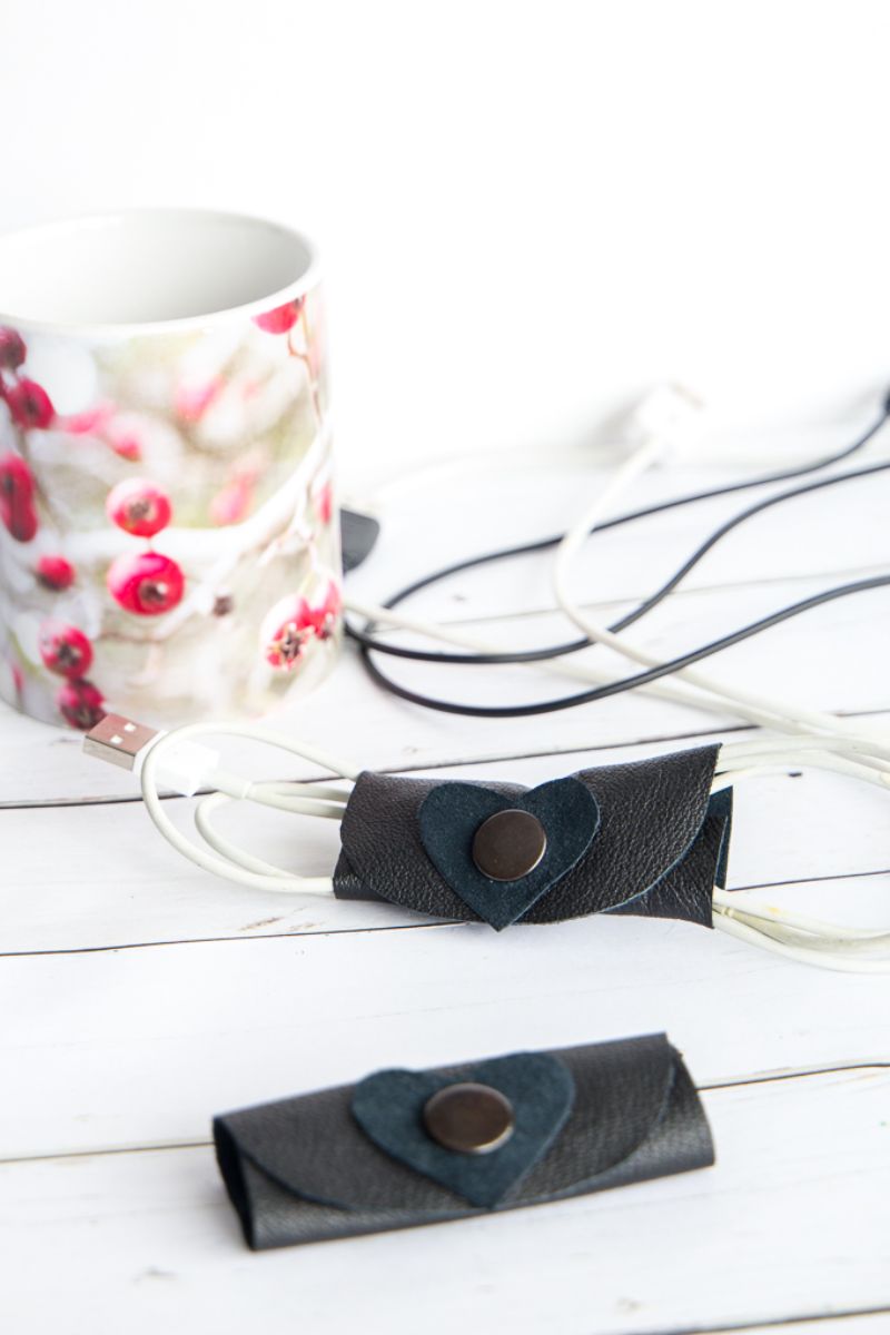 A white mug with a red berry design sits on a white wooden surface. Beside it are black leather cord organizers with snap buttons and heart-shaped adornments, neatly binding white and black cables. Another cord organizer lies nearby, not in use—perfect small gifts to mail to friends.