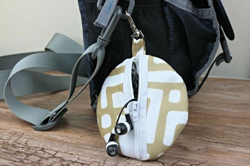 Small, round zippered pouch with a beige and white geometric pattern attached to a gray backpack. The pouch is partially open, revealing a black pair of earphones with silicone tips hanging out. Ideal for small gifts to mail to friends, the backpack has mesh pockets and adjustable gray straps.