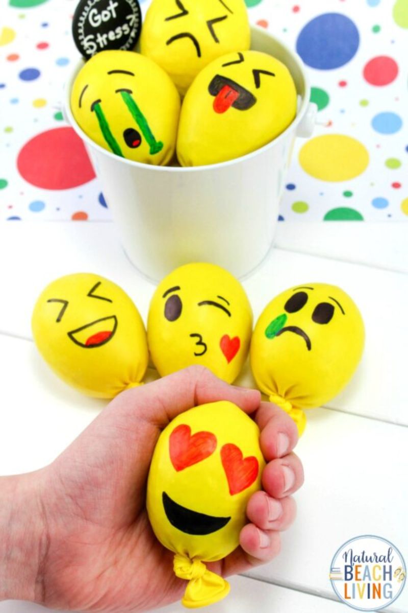 A hand holds yellow stress balls decorated with various emoji faces, including a smiling face with heart eyes, while other emoji balls rest on a table and in a white container in the background. The table is sprinkled with colorful confetti, making for perfect small gifts to mail to friends.