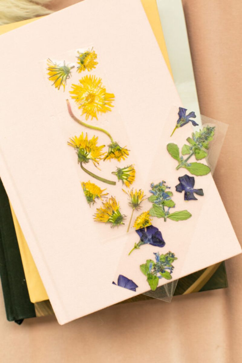A pink notebook with pressed yellow and purple flowers arranged on its cover rests on top of other books with various colored covers, all placed on a light tan surface—a perfect small gift to mail to friends.