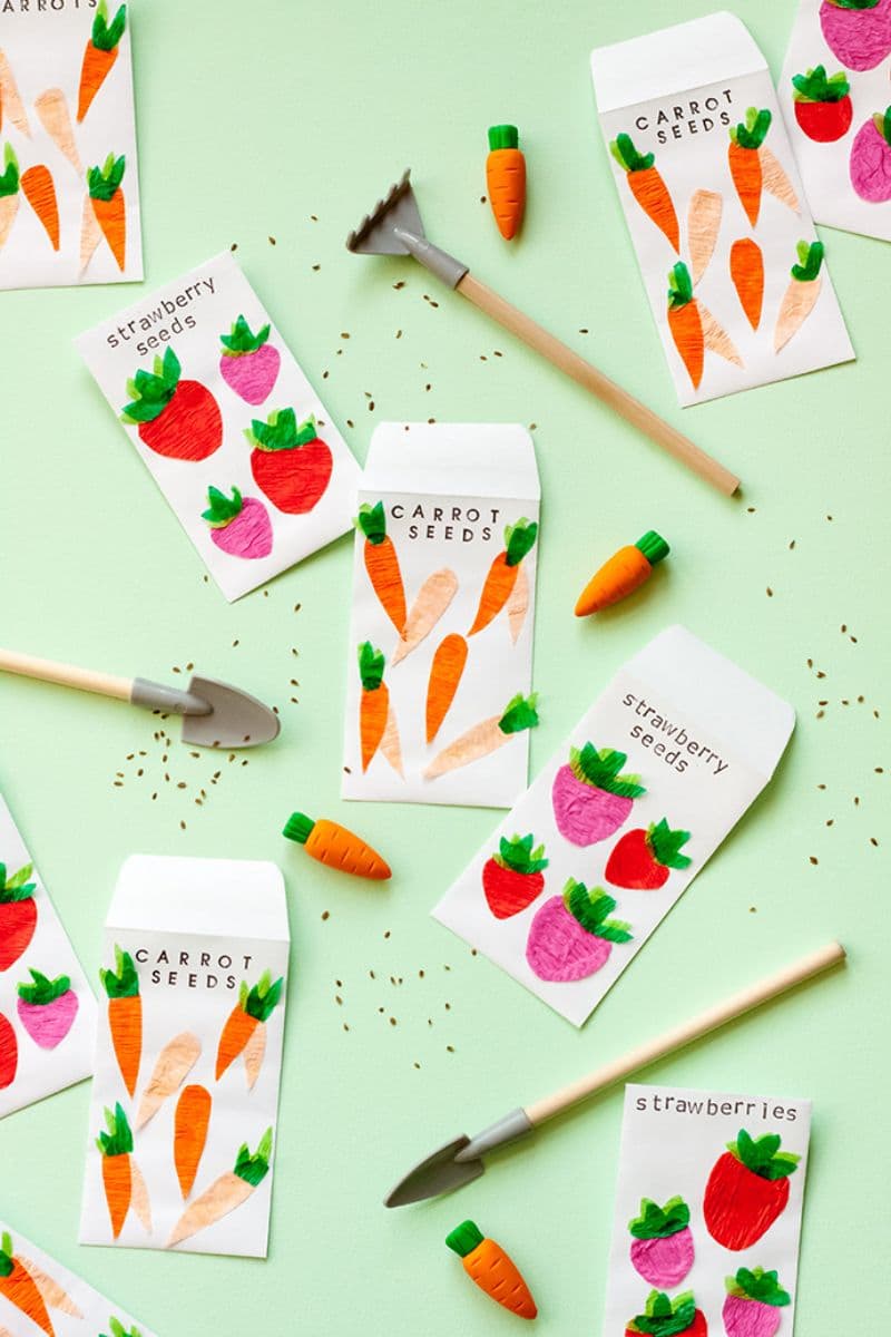 Seed packets labeled "Carrot Seeds" and "Strawberry Seeds" are scattered on a light green surface. Miniature garden tools and small carrot-shaped objects are arranged among the packets, along with scattered seeds. These small gifts make perfect surprises for friends in your next mail.