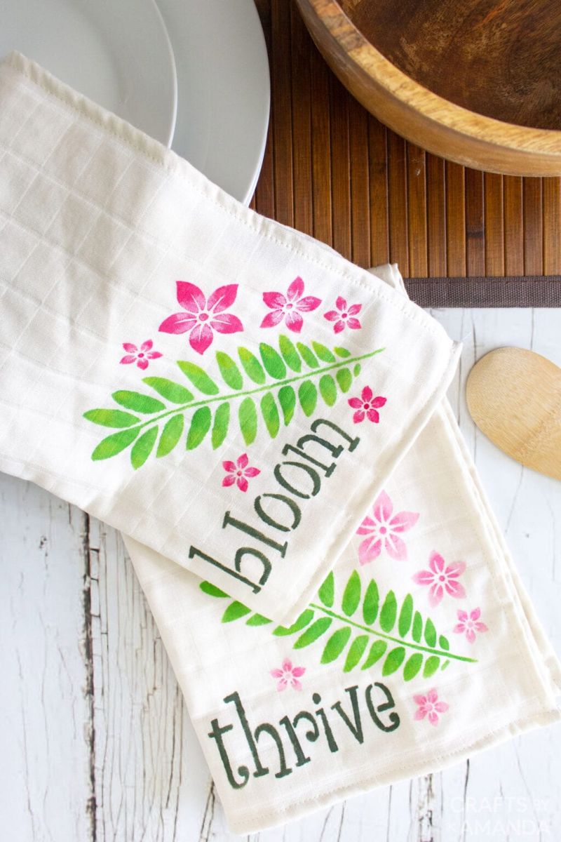 Two white kitchen towels are decorated with floral-themed designs, perfect for simple gifts or Mother’s Day gifts. One towel features the word "bloom" with pink flowers and green leaves, and the other showcases the word "thrive" with similar decorations. Plates and a wooden bowl are partially visible.