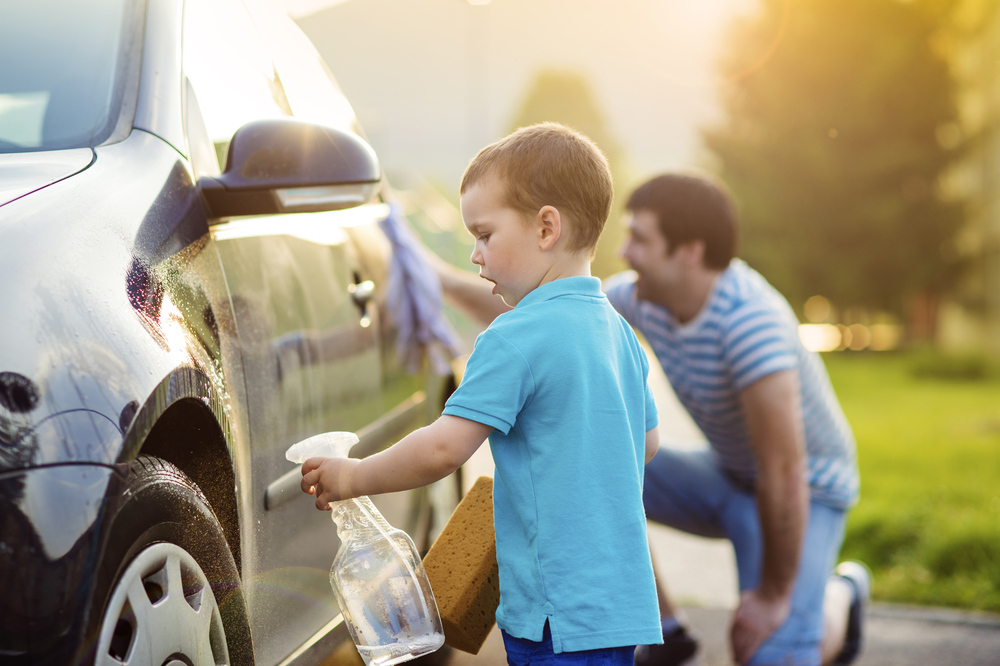 A young boy sprays water on a car he's washing, holding a sponge in his other hand. In the background, an adult man washes the car's tires with a cloth. They are outdoors on a sunny day, working together as part of their Mother's Day surprise ideas.