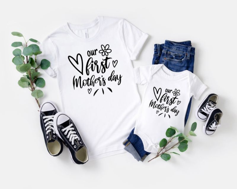 A flat lay of matching outfits for a mother and baby, featuring a white t-shirt and a white onesie with the text "our first Mother's day" and heart designs. Perfect for new moms, the set is paired with black sneakers, baby shoes, jeans, and leafy green branches. Ideal Mother's Day gifts!