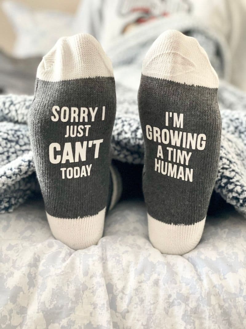 A pair of feet with gray and white socks laying on a bed with a gray blanket. The left sock reads "SORRY I JUST CAN'T TODAY" and the right sock reads "I'M GROWING A TINY HUMAN," making it perfect for expecting mothers or new moms as Mother’s Day gifts.