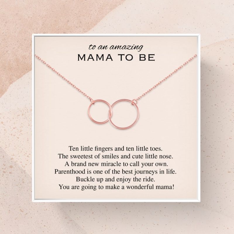 A square card featuring a rose gold necklace with two interlocking circles. The card is addressed "to an amazing Mama to Be" and includes a heartfelt message for expecting mothers about the joys of parenthood, mentioning baby toes, smiles, and the excitement of welcoming a new miracle.