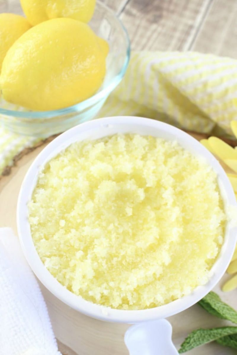 A small white bowl filled with bright yellow lemon sugar scrub sits on a wooden surface, making it one of the perfect simple gifts. Next to it, a glass bowl holds whole lemons. A folded yellow and white striped towel and a few green leaves are also visible nearby, suggesting an ideal DIY Mother’s Day project.