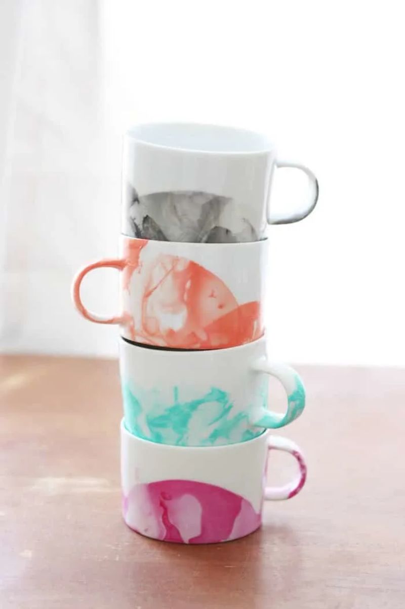 Four white ceramic mugs are stacked vertically on a wooden surface, perfect for Mother’s Day gifts. Each mug features a unique, colorful marbled design: the top in shades of gray, the second in orange, the third in teal, and the bottom in pink. Natural light filters in from the background.