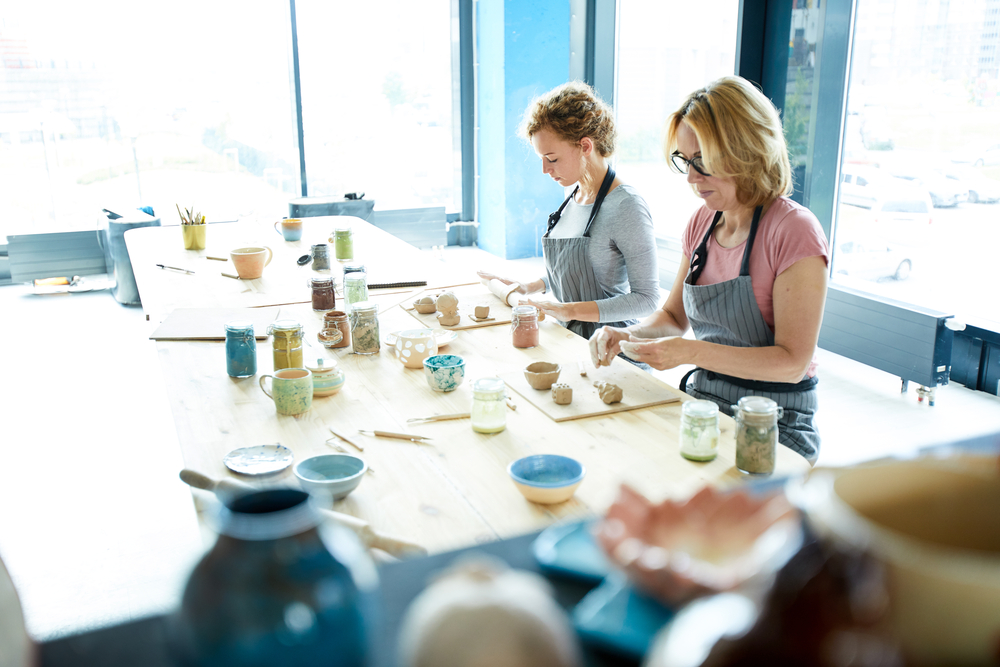 Two women wearing aprons are sitting at a table, engaged in pottery making. The table is filled with ceramic tools, various pottery items, and jars of paint. Bright natural light streams through large windows in the background—a perfect scene for Mother's Day surprise ideas.