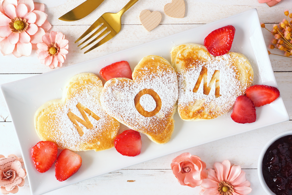 A rectangular white plate holds three heart-shaped pancakes dusted with powdered sugar. The middle pancake has "MOM" spelled out with cut-out letters. Sliced strawberries and pink flowers decorate the plate. Golden cutlery and wooden heart decorations are in the background, perfect for Mother's Day surprise ideas.
